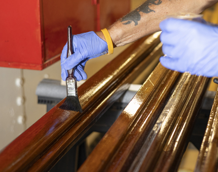 A close up of a person wearing blue gloves holding a paintbrush, adding a layer of varnish to a handrail. 