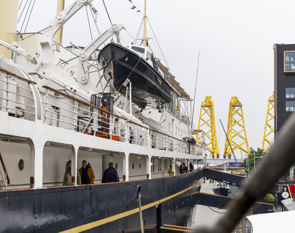A view of the side of Britannia, there are people walking on the deck, in the background there are tall yellow structures in the Port of Leith.