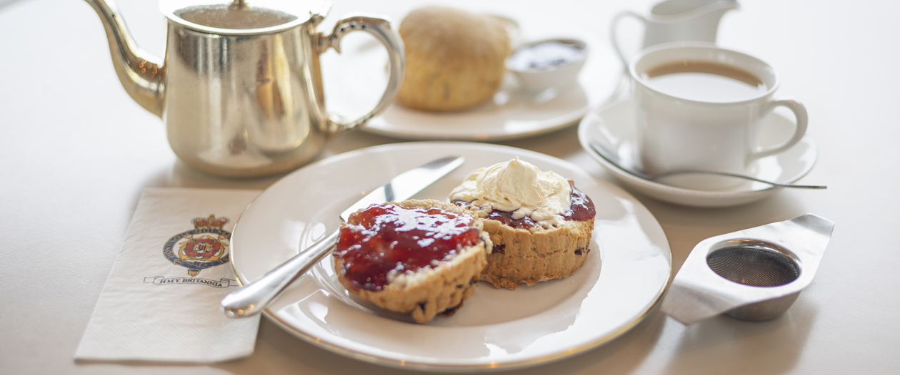  A traditional cream tea is set on a white tablecloth with a silver teapot, a cup of tea, a small milk jug, and two scones topped with jam and clotted cream. A napkin printed with the Britannia crest sits beside the plate.