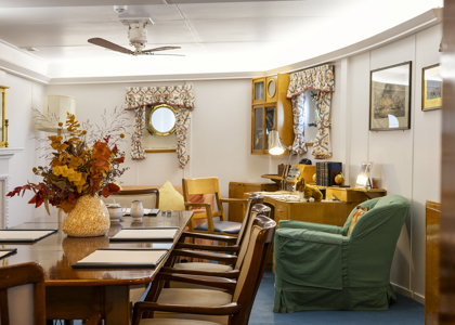 A warm, wood‑panelled room with nautical touches, featuring a large table with chairs, a desk with a lamp, a green armchair, and two porthole windows with patterned curtains. A vase of autumn‑coloured flowers sits on the table, adding to the cosy, lived‑in feel.