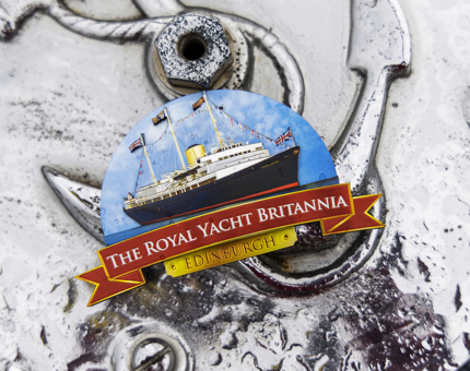 A magnet with an illustration of Britannia on it, it also has a red ribbon design at the bottom with The Royal Yacht Britannia written on it. 