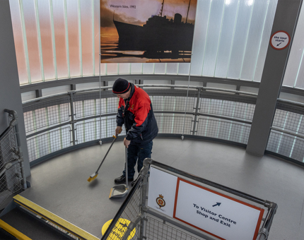 A Facilities Officer is in the stairwell at The Royal Yacht Britannia with a broom and dustpan sweeping. 