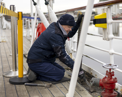 A Maintenance man kneeling next to the railings aboard Britannia, he's scraping dirt from the decks. 