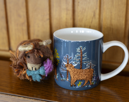 A mug with an illustration of a Highland cow and a small toy of a Highland cow are sitting on a wooden shelf. 