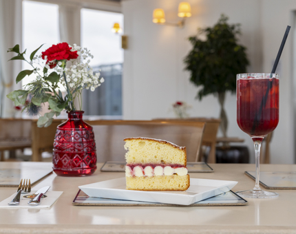 A red coloured cocktail and slice of Victoria sponge cake are on a table with a small vase holding a red rose and baby's breath flowers. 