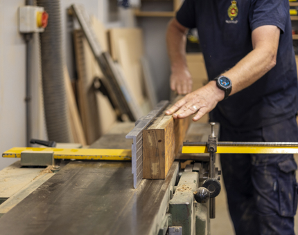 A Maintenance team member is at a work bench, planing a piece of wood. 