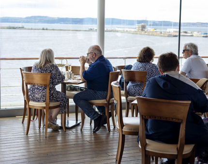 Guests sitting at tables in the Royal Deck Tearoom. Through the big windows you can see the water of the Port of Leith in Edinburgh. 