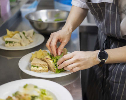 A Chef is in the Galley aboard Britannia in Leith. They are adding salad to plates of sandwiches. 