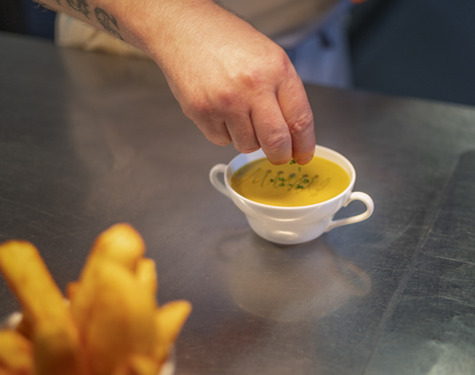 In the Galley, a hand sprinkling chives into a bowl of soup, there is a bowl of potato wedges in the foreground. 