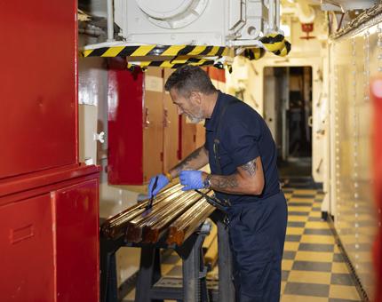 A Maintenance man is in a hallway with red walls and lack and white checkered floor, he is varnishing handrails. 