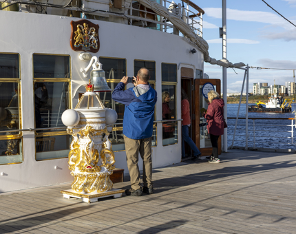 A man is on the Verandah deck standing next to the decorative binnacle and taking a picture of Britannia's Bell. In the background you can see the water with a boat in the Port of Leith. 