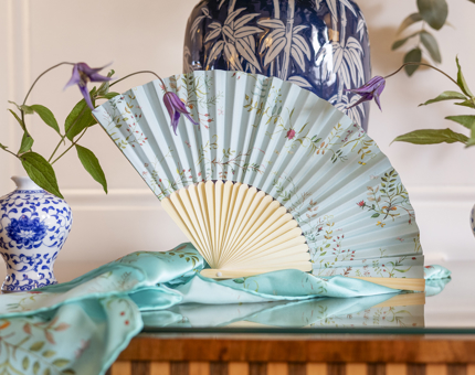 A fan and silk scarf from the Shop's Embroidery Range is on a table with vases of flowers. 