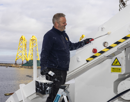 A maintenance man is holding a paint bucket, paint brush and roller, applying a coat of white paint. The Port of Leith is in the background. 