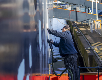 A Maintenance team member grinding down patches of paint on Britannia's hull. 
