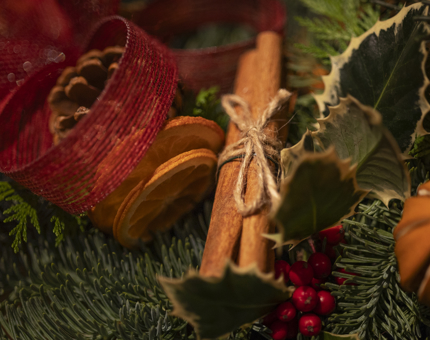 A close-up of foliage used to make a wreath, there is cinnamon sticks, dried orange slices, red ribbon and pine cones. 