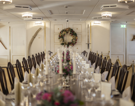 The long dining table in the State Dining Room is set. There is a Christmas wreath hanging on the wall at the back of the room. 