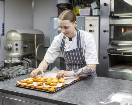 A chef is in the Galley, she is at the bench putting almond financiers on a tray. 