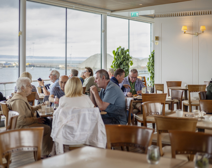 Visitors sitting at tabled in the Royal Deck Tearoom. In the background there are ceiling height windows looking out to the Port of Leith.