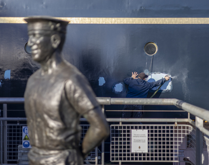 A Statue of Norrie, the longest serving Yachtsmen on the Quayside. In the background a Maintenance team member is grinding paint down on the hull of Britannia. 