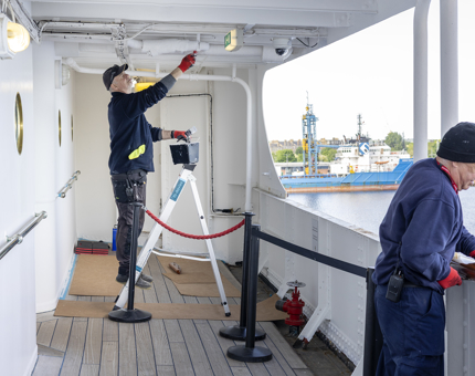 A man is standing on a ladder painting the ceiling of a deck on board Britannia. 