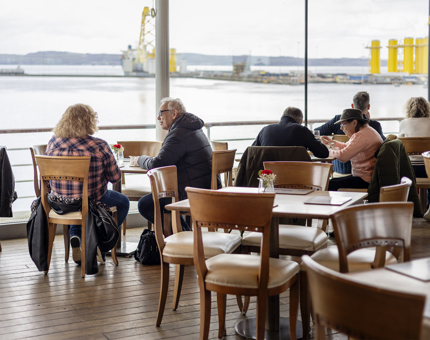 Visitors are sitting at tables in the Tearoom aboard Britannia. There are large windows where you can see the Port of Leith through. 