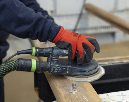 A close up of a hand wearing a red protective glove holding an electric sander to sand a wooden handrail. 