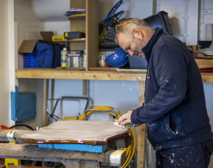 A Maintenance team member is sanding a wooden crest in the workshop. 