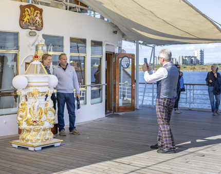 A Visitor Assistant is taking a photo of visitors at the Bell aboard Britannia in the Port of Leith. 
