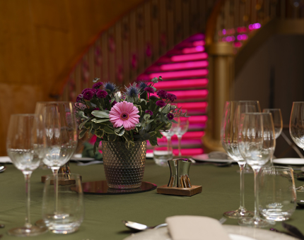 A close up of a table set for an event in Fingal's Ballroom. The table cloth is green and there is a vase with pink and purple flowers. 