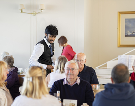 Busy tables in the Tearoom, a waiter is serving people who are seated. 