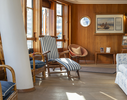 The Sun Lounge aboard The Royal Yacht Britannia. It is wood paneled and has a couch and a stripy blue lounge chair.