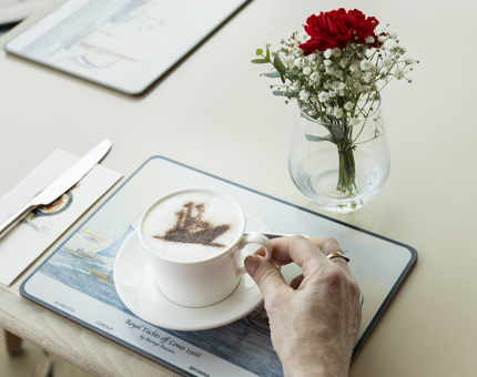 A hand holding a cappuccino with a chocolate stencil of a ship on it. 