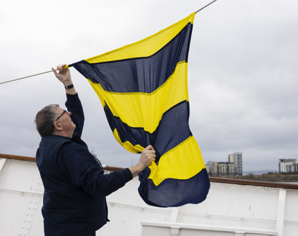 A man is raising a blue and yellow dress flag above Britannia in Leith. 