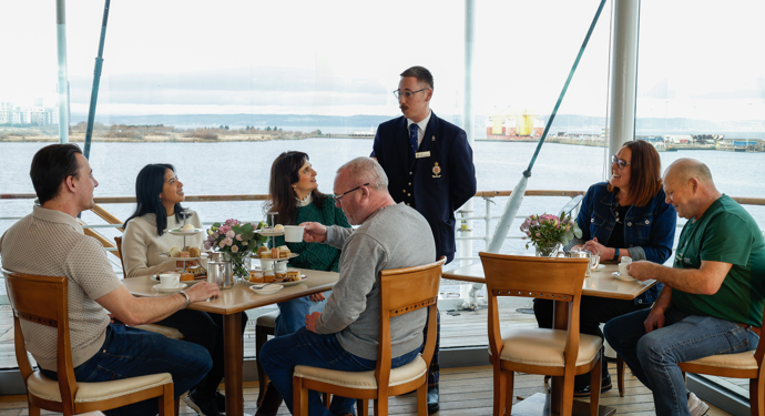 Guests Dining In The Royal Deck Tearoom Aboard The Royal Yacht Britannia. A tour guide is speaking to them. 