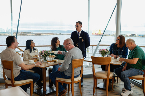 Guests Dining In The Royal Deck Tearoom Aboard The Royal Yacht Britannia. A tour guide is speaking to them. 