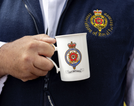 A close up of a hand holding a white china mug with a colourful Britannia crest on it. 
