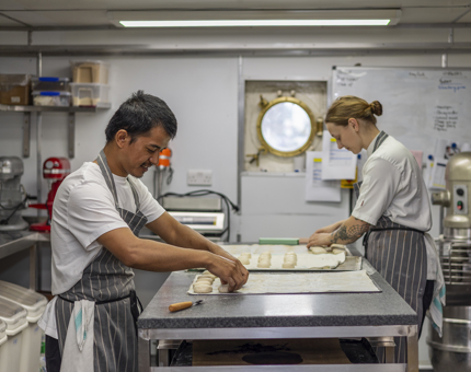 Two chefs are in the Galley standing at a bench making bread rolls.