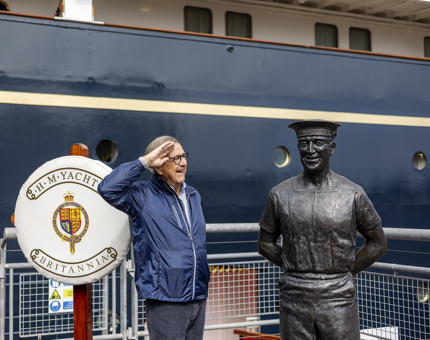 A man stands next to a statue of a sailor and salutes him at The Royal Yacht Britannia in Edinburgh.