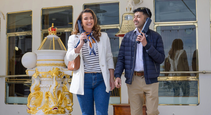 A man and woman on the Verandah Deck of The Royal Yacht Britannia listening to audio guide handsets.