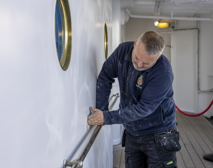 A man from the Maintenance team is sanding a wall on the ship. 