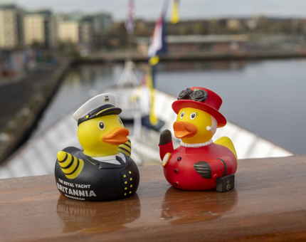 An Admiral and Queen rubber duck positioned on a wooden railing with the Port of Leith in the background. 